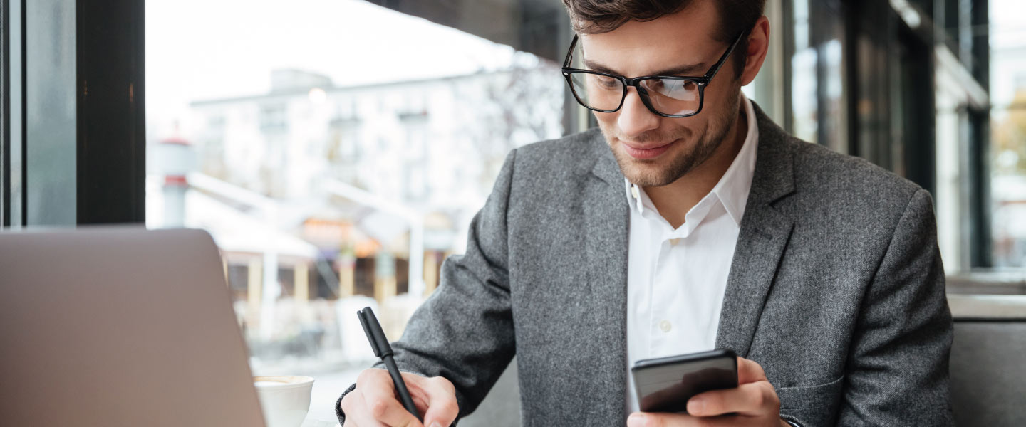 Empresário sorridente em óculos, sentado junto à mesa no café com o computador portátil enquanto estiver usando o smartphone e escrevendo algo