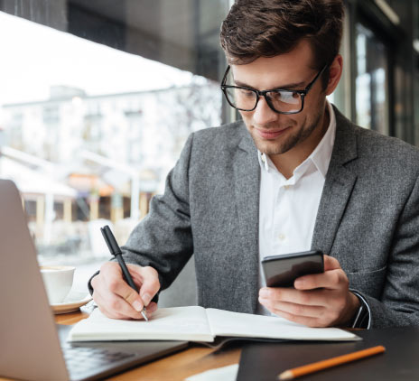 Empresário sorridente em óculos, sentado junto à mesa no café com o computador portátil enquanto estiver usando o smartphone e escrevendo algo